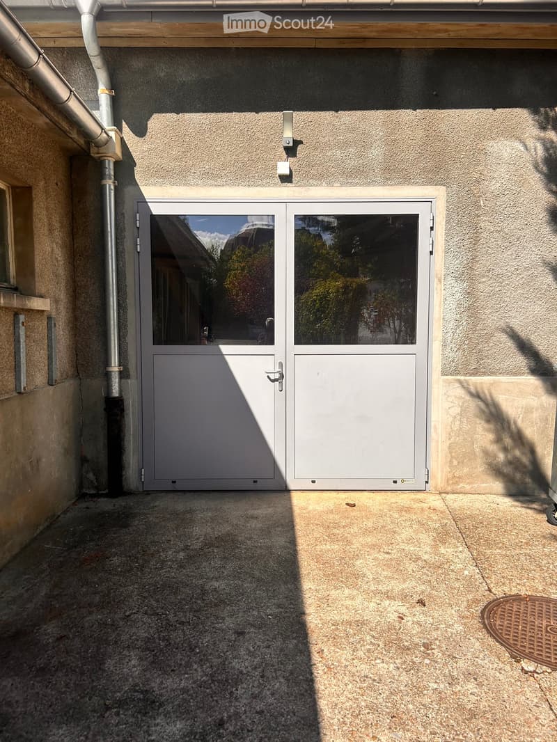 A narrow concrete pathway leads to a white double door with glass panels, flanked by textured beige walls and a metal pipe on the left side.