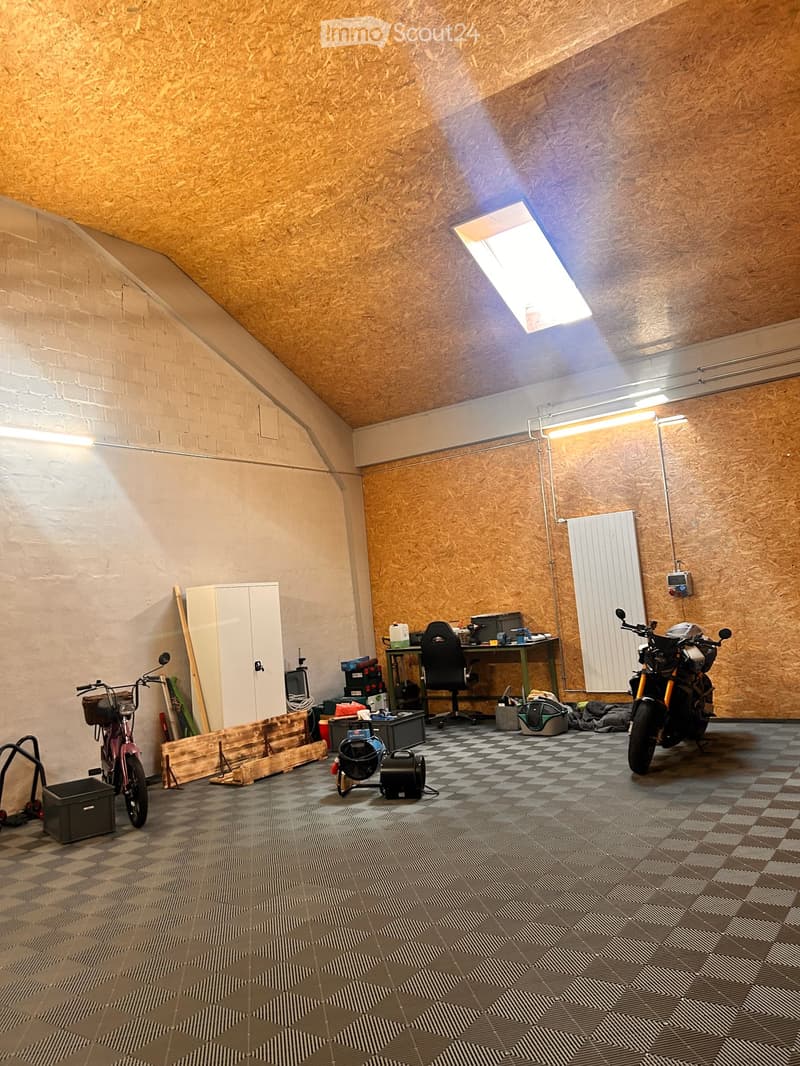 A spacious workshop or garage with a slanted wooden ceiling, exposed brick wall, and checkered floor. Contains a desk with a chair, a motorcycle, a bicycle, a fan, a radiator, and various storage items.