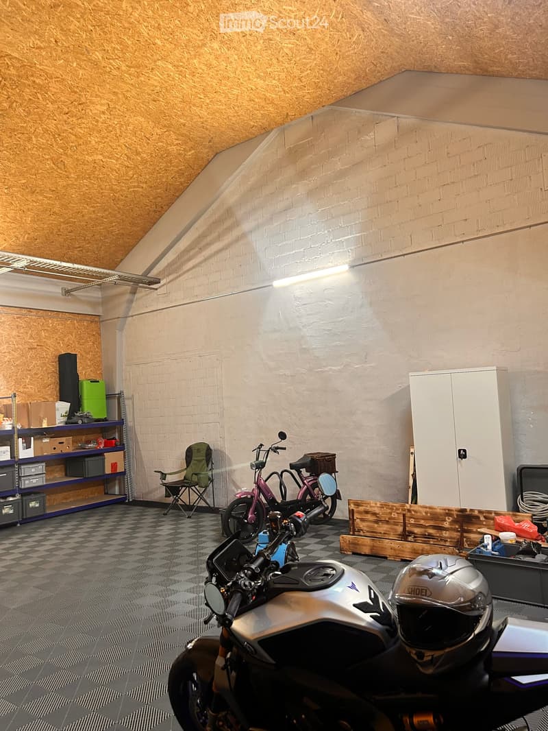 Garage with a slanted wooden ceiling, white brick walls, and a checkered floor. Contains a motorcycle, a helmet, a folding chair, shelves with storage boxes, and a wooden plank on the floor.