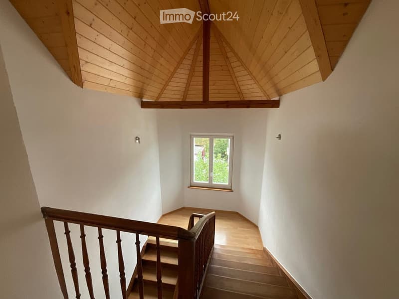 Staircase with wooden steps and handrail, white walls, and a small window at the top showing greenery outside.