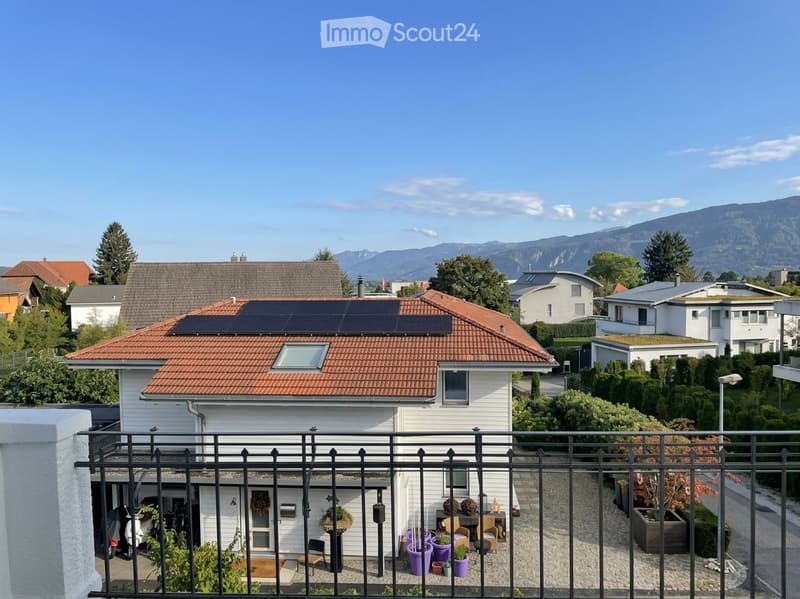 A two-story house with a red-tiled roof, white exterior, and solar panels on the roof. The house features a small balcony area with potted plants and outdoor furniture.
