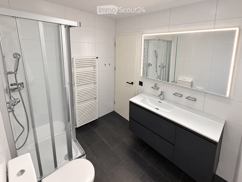 Modern bathroom with a white tiled shower area, a black vanity with a sink and mirror, a toilet, and a radiator. The floor is dark grey tiles.