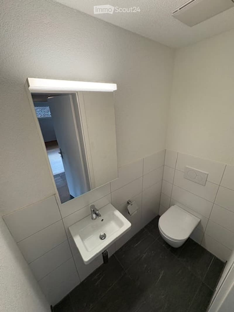 Modern bathroom with white tiled walls, dark floor, sink, toilet, and mirror with built-in lighting.