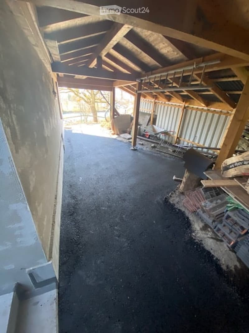 A covered outdoor corridor with a concrete floor, wooden beams, and a partially constructed wall on one side. The area appears to be under construction or renovation, with some tools and materials visible.