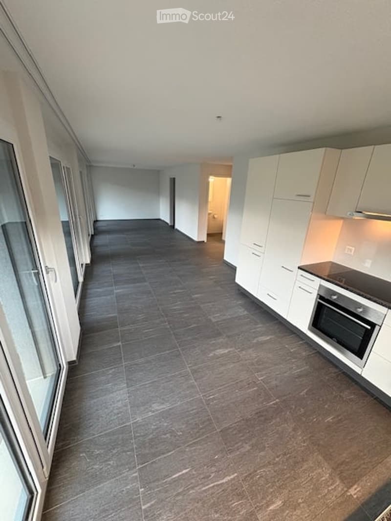 Empty kitchen with white cabinets, dark tiled floor, and large windows. No furniture or appliances visible.