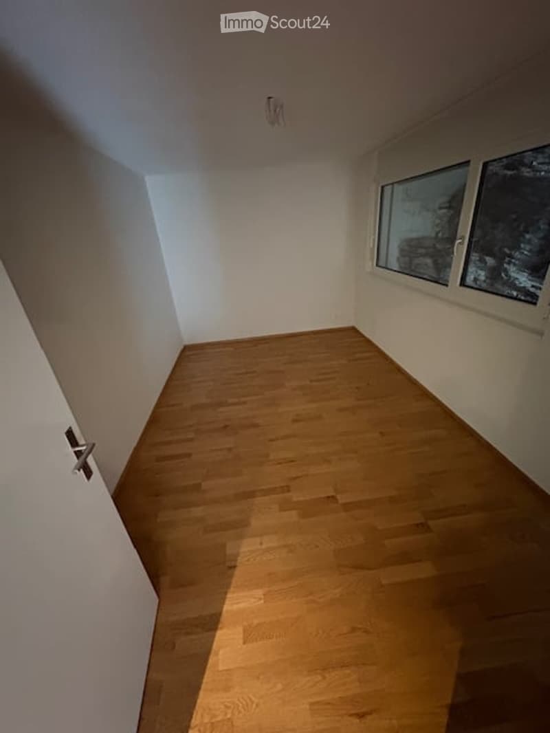 Empty room with wooden flooring and white walls, featuring a door on the left and windows on the right.