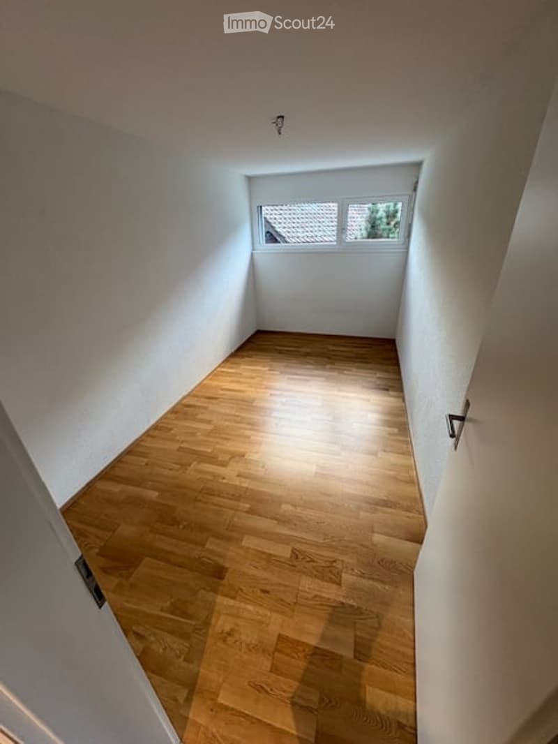 Empty hallway with wooden flooring and white walls, featuring two windows at the far end and a door on the right side.