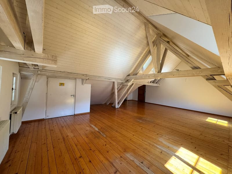 An empty attic space with wooden floors, exposed wooden beams, and skylights allowing natural light to enter. The room features a staircase leading to another area and a closed door.