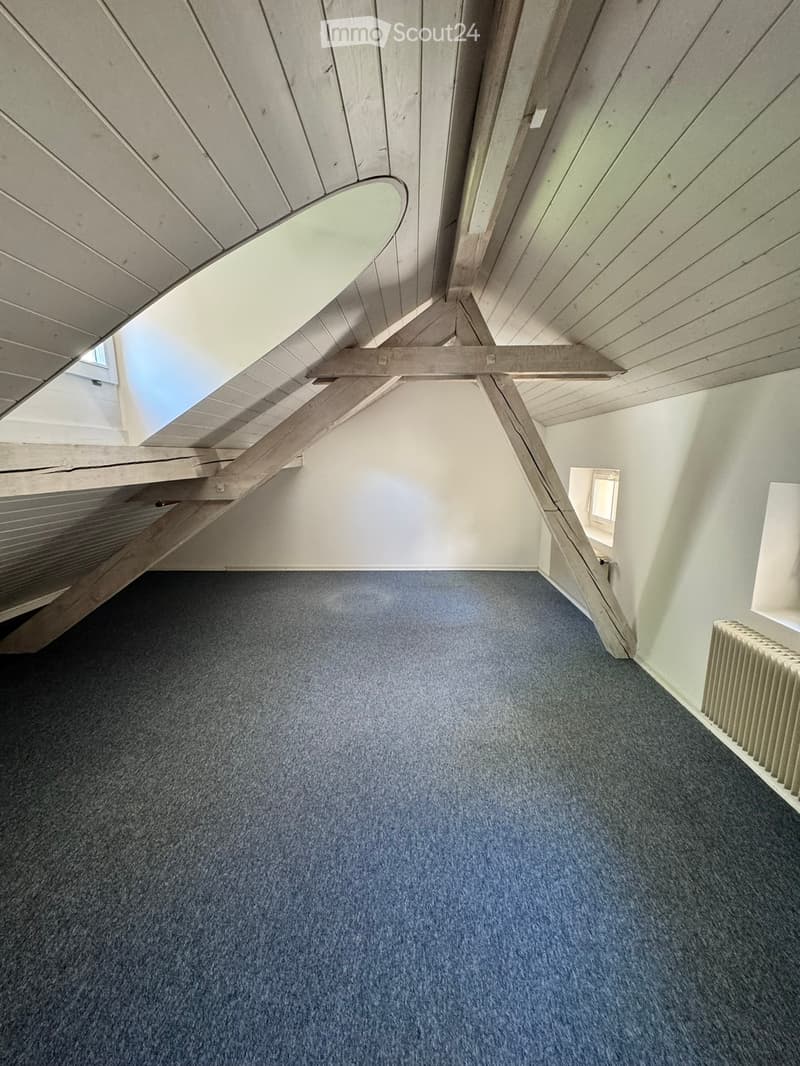 Empty attic space with exposed wooden beams, blue carpet, and small windows allowing natural light.