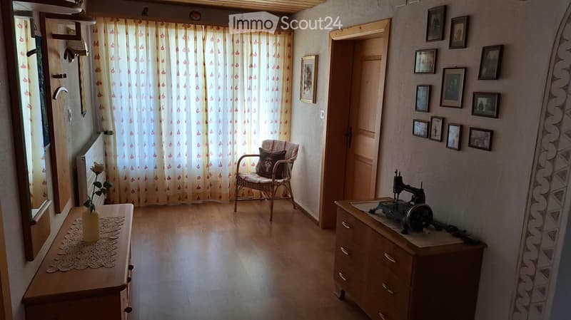 A cozy, well-lit room with wooden flooring, a wicker chair with a cushion, a small table with a flower vase, a wooden dresser with a sewing machine, and several picture frames on the wall.