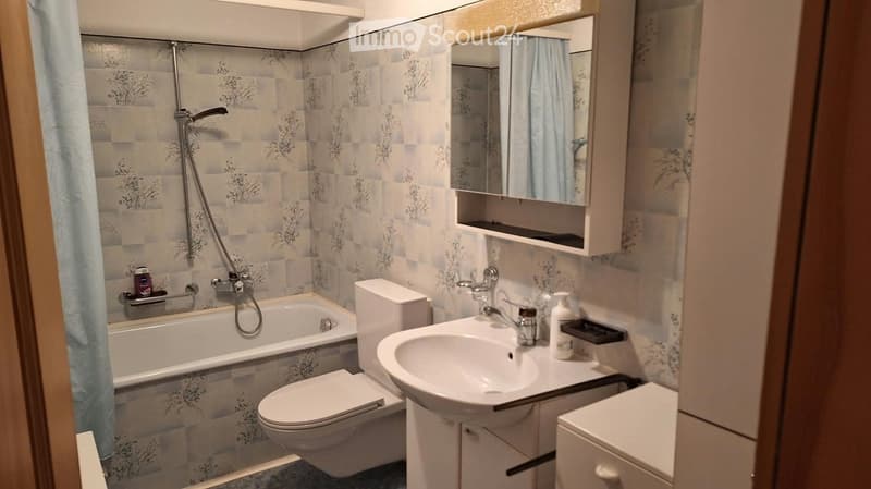 Bathroom with a bathtub, toilet, sink, mirror, and shower hose. The walls are tiled with a floral pattern, and there is a cabinet above the sink.