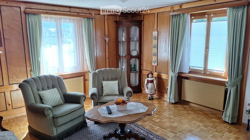 Living room with wooden walls, hardwood floor, large windows with curtains, two armchairs, coffee table with a table runner and remote control, a wooden cabinet, and a decorative doll.