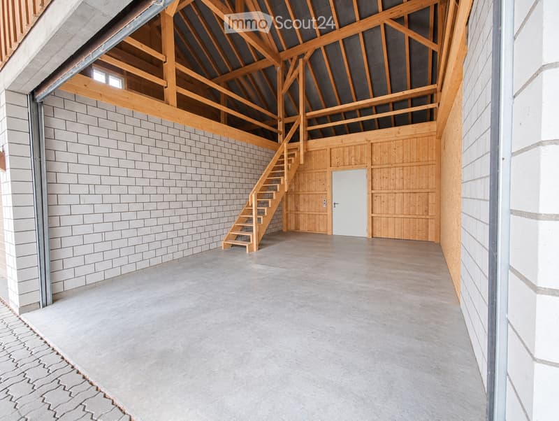 An empty, spacious room with exposed wooden beams and a high ceiling, featuring a staircase leading to an upper level, white brick walls, and a concrete floor.