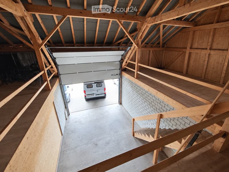 A spacious, unfinished garage with exposed wooden beams and a concrete floor, featuring a large garage door and a staircase leading to an upper level.