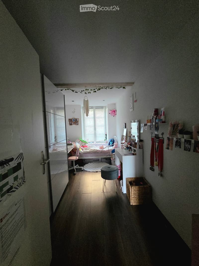 A narrow hallway leads to a bedroom with a bed, nightstand, and various decorations. The hallway has wooden flooring and walls with artwork and shelves.