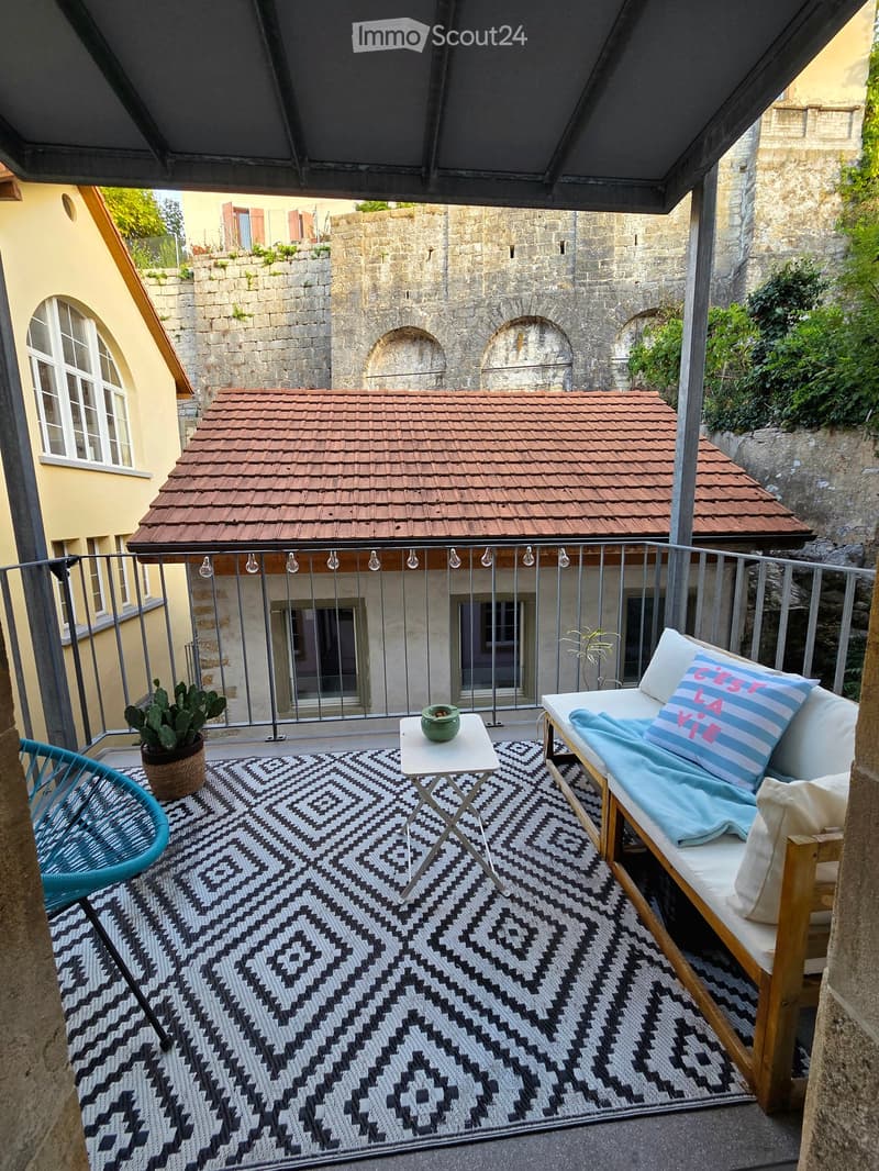 Balcony with a patterned rug, a small white table, a blue sofa with cushions, a turquoise chair, and a potted plant.