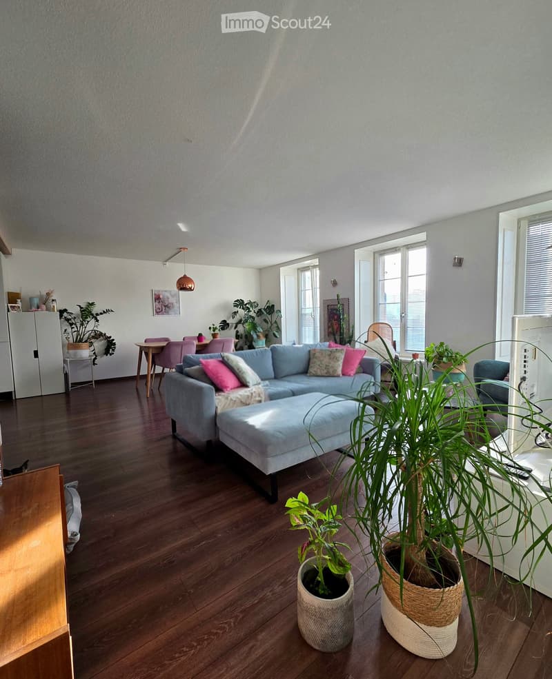 Bright living room with wooden flooring, large windows, a blue sectional sofa with pink and floral cushions, a matching ottoman, a small dining area with a table and chairs, and several potted plants.