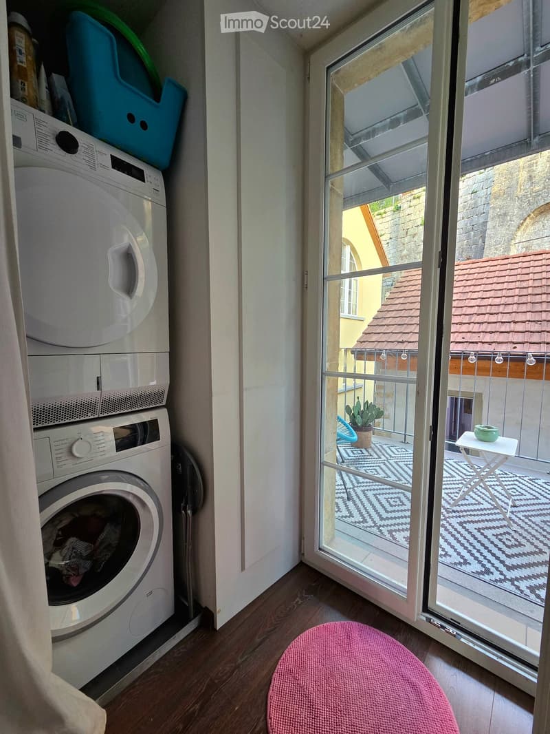 A small room with a washing machine and a pink stool, leading to a balcony with a small table and a potted plant.