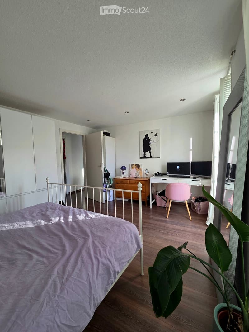 A well-lit bedroom with a wooden floor, a bed with a purple duvet, a desk with a computer, a pink chair, a plant, and a mirror on the wall.