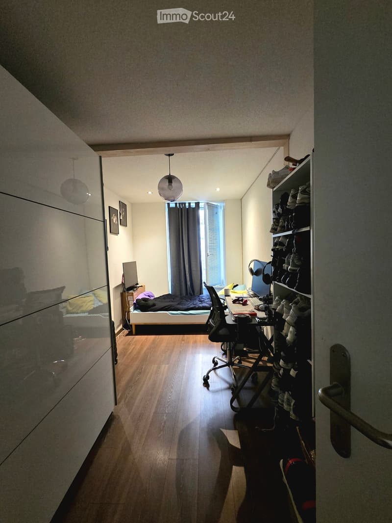 A furnished bedroom with a bed, desk, chair, screen, shelf, and headphones. The room has wooden flooring and a window with curtains.