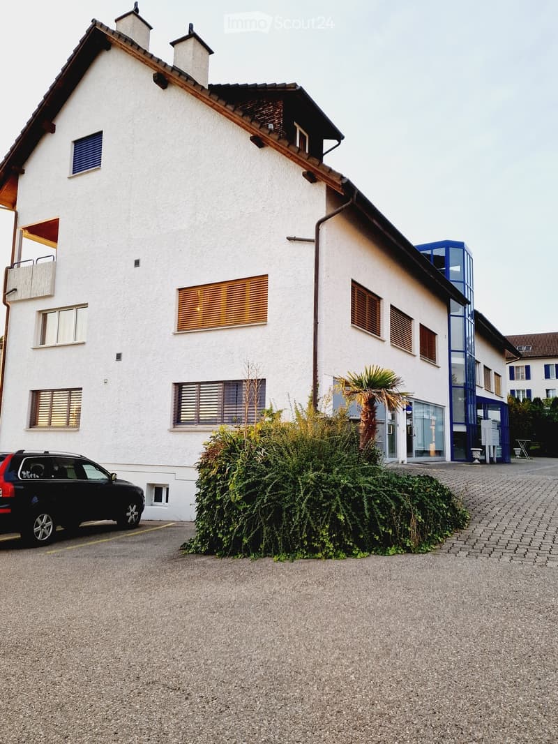 Two-story white building with multiple windows, some with shutters, and a small balcony. A black SUV is parked in front, and there is a small garden area with a palm tree and shrubs.