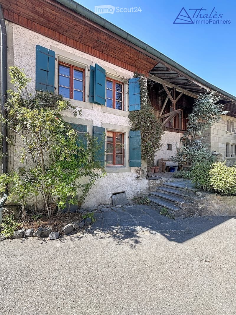 Two-story stone house with wooden shutters, a covered wooden porch, and a gravel driveway.