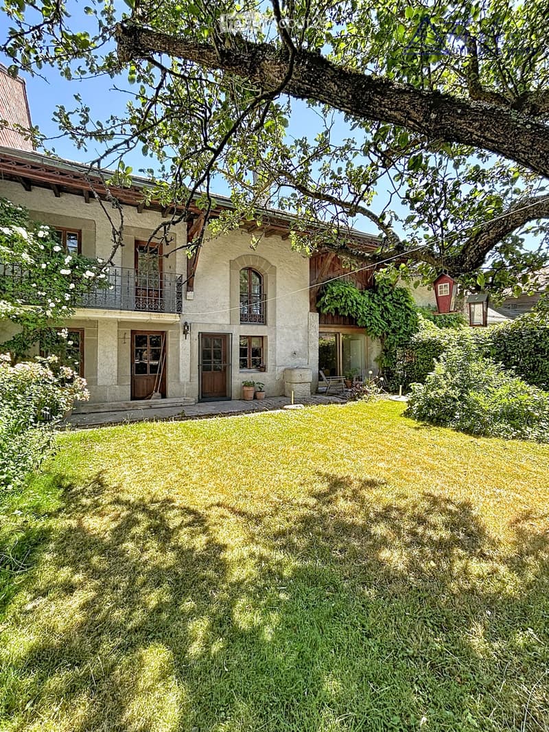 A two-story house with beige stone walls, wooden doors and windows, a balcony with metal railings, and a well-maintained lawn.