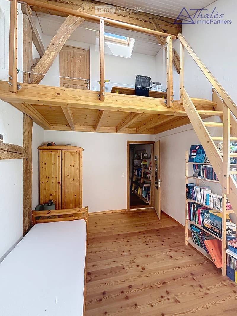 Cozy, wooden-floored room with a lofted workspace area, a bed, bookshelf, and cabinet.