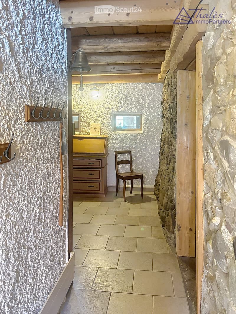 Narrow hallway with stone walls, wooden ceiling beams, tiled floor, small window, wooden chair, and a small wooden cabinet with drawers.