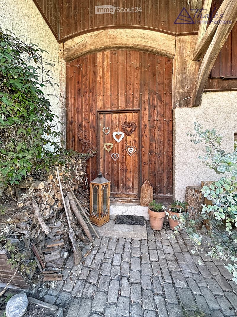 Entrance to a rustic building with a wooden door adorned with heart-shaped decorations, a lantern, potted plants, and a cobblestone pathway.