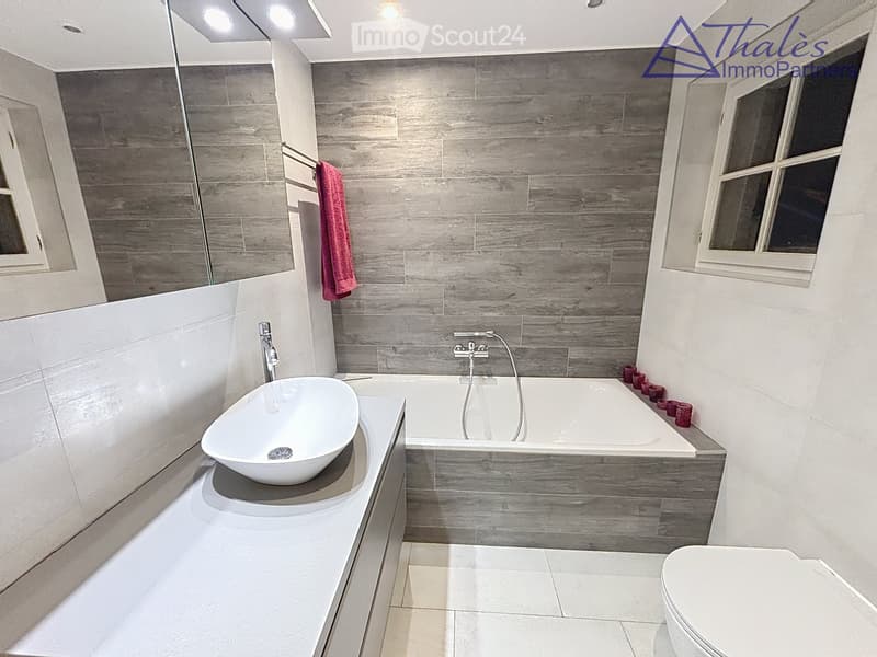 Modern bathroom with a large bathtub, a white sink, a toilet, and a towel hanging on the wall. The walls are tiled in a wood-like pattern, and the floor is tiled in white.