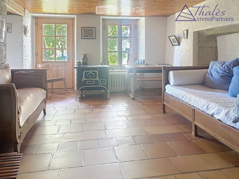 Cozy living room with tiled floor, stone walls, and wooden ceiling beams. Features a sofa, armchair, small table, and radiator.