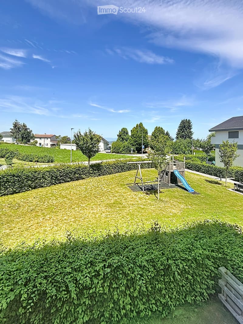 A well-maintained garden with a swing set, slide, and surrounding greenery, bordered by hedges and trees.