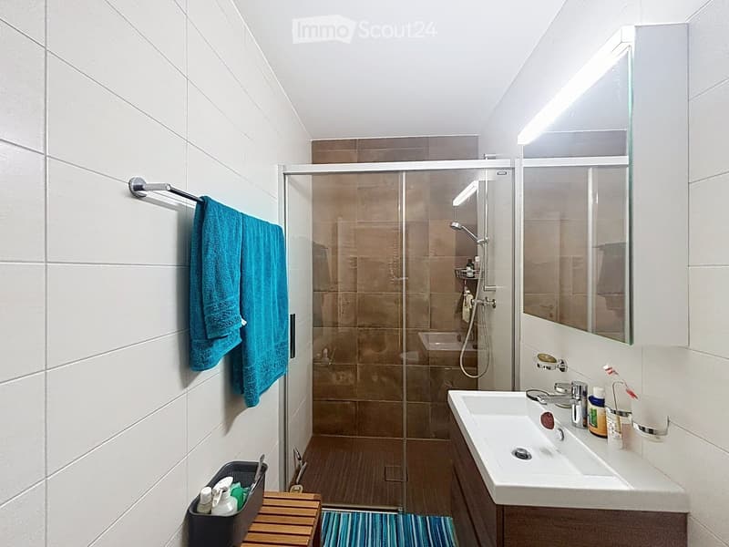 Modern bathroom with white tiles, a glass-enclosed shower, a large mirror, a sink with a cabinet, and a towel hanging on a rack.