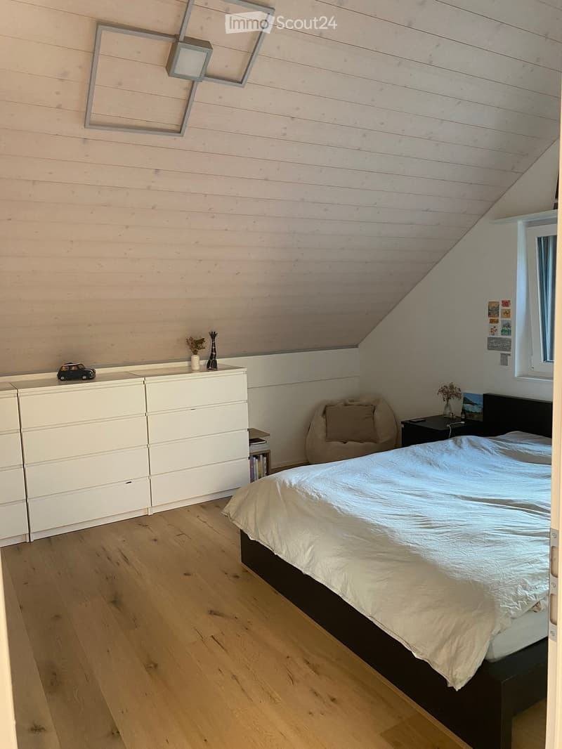 Modern bedroom with wooden flooring, slanted ceiling, white walls, and built-in storage. Features a large bed with white bedding, a nightstand with a lamp, and a drawer unit with decorative items.