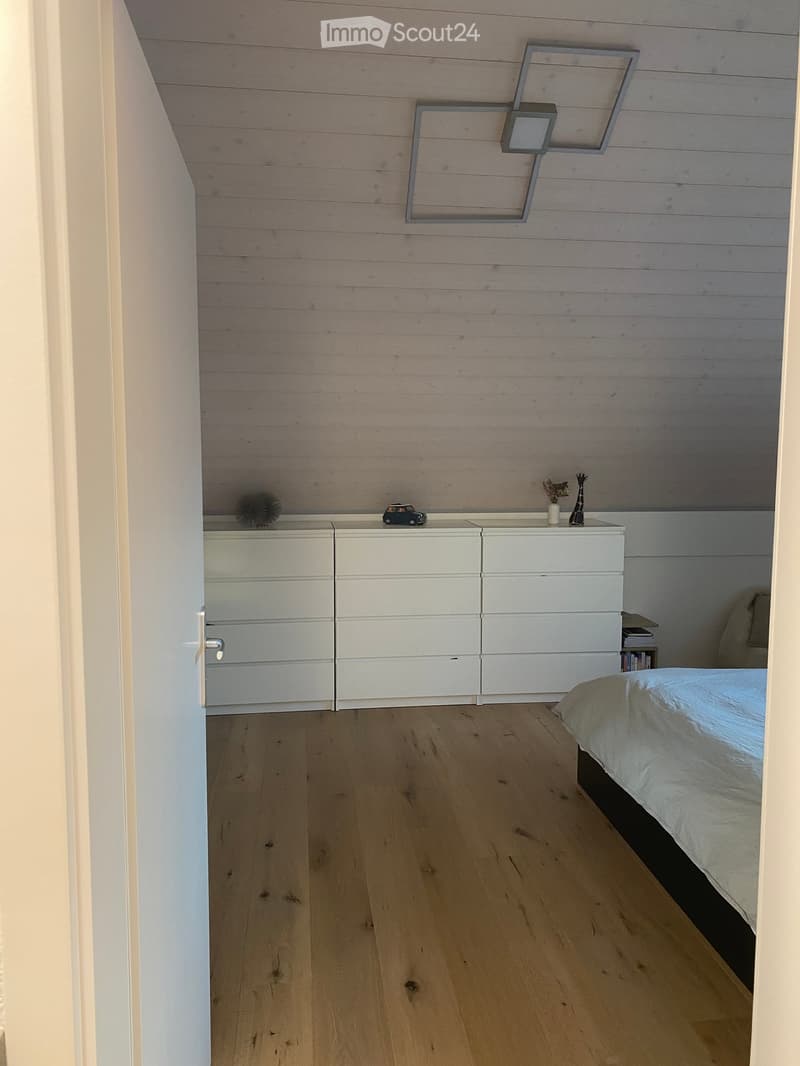 Modern bedroom with light wooden flooring, white nightstand, and a bed with white bedding. The walls are paneled in light wood, and there is a decorative wall-mounted light fixture above the nightstand.