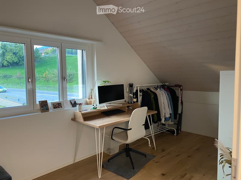 A modern, well-lit attic workspace with a wooden desk, ergonomic chair, computer setup, and a clothing rack. Large windows offer a view of the garden and road.