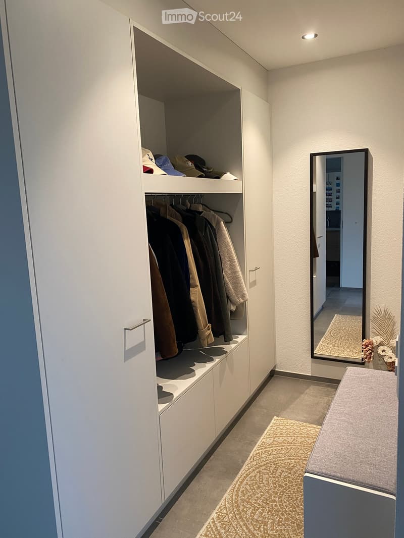 Modern hallway with built-in white closet, clothing items, hats, and a large mirror reflecting a small room with a rug and decorative items.