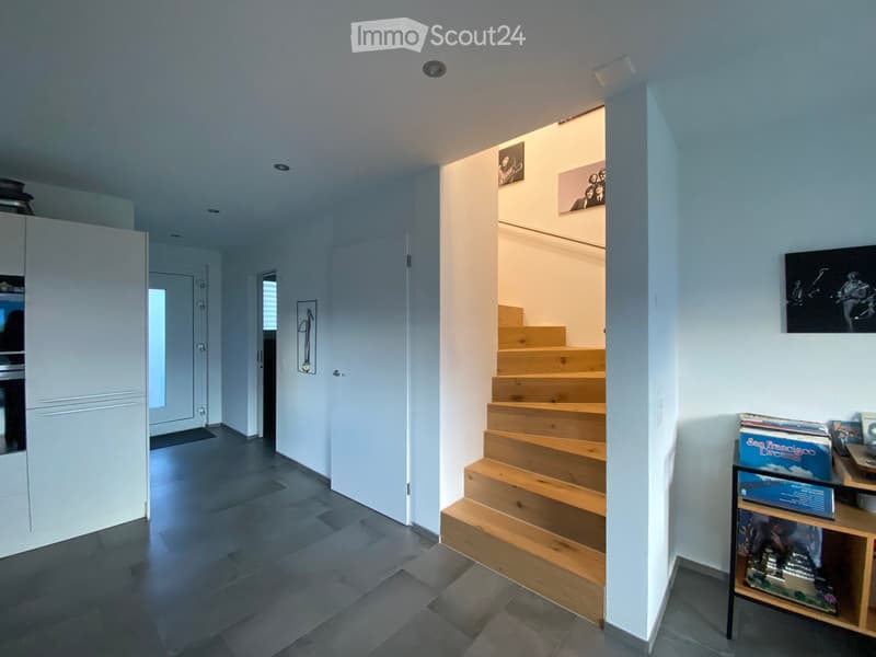 Modern hallway with grey tiled floor, white walls, wooden staircase leading upwards, and a small shelf with books and records on the right side.