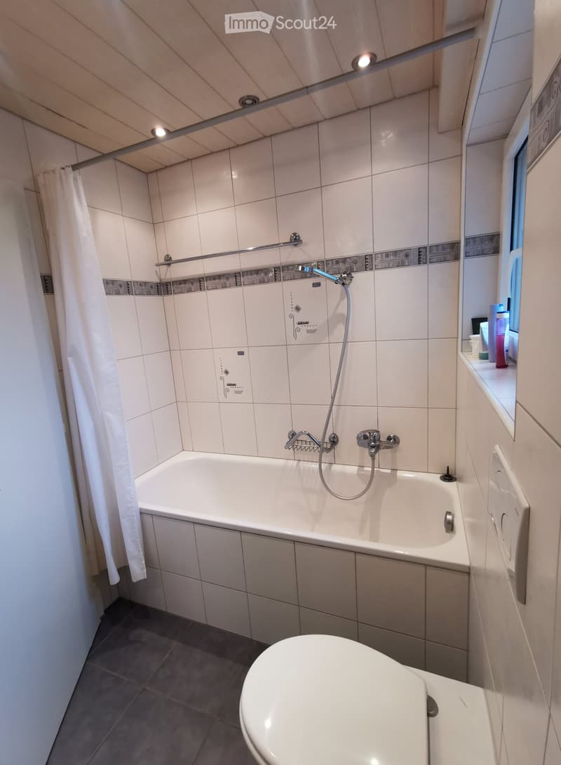 Modern bathroom with a bathtub, toilet, shower, and a window with a small shelf holding toiletries.