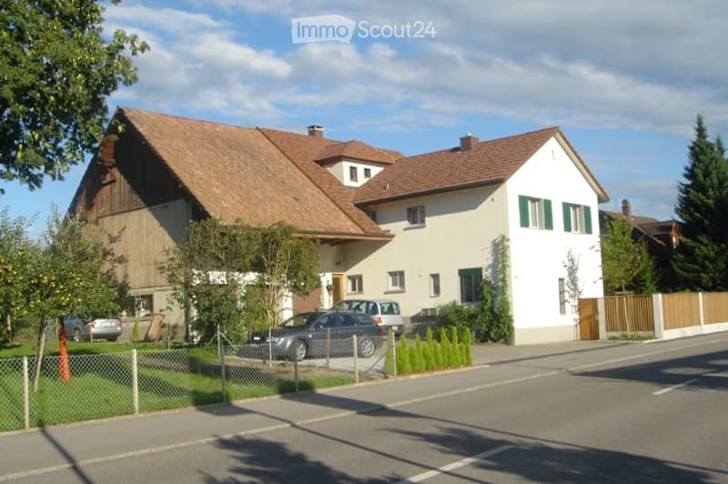 Two-story house with a red-tiled roof, white walls, green shutters, and a fenced front yard with parked cars.
