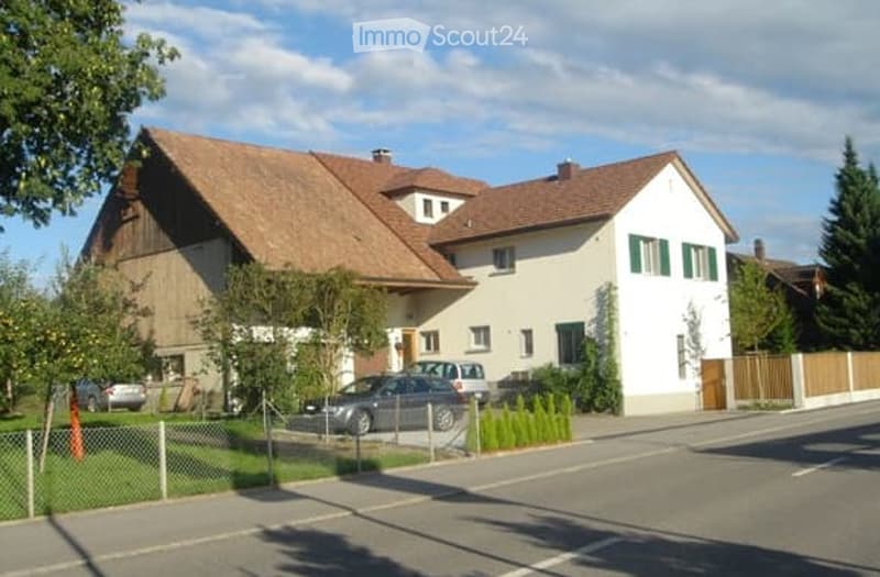 Two-story house with a red-tiled roof, white walls, and green shutters. A car is parked in the driveway, and there is a small garden area with a fence.