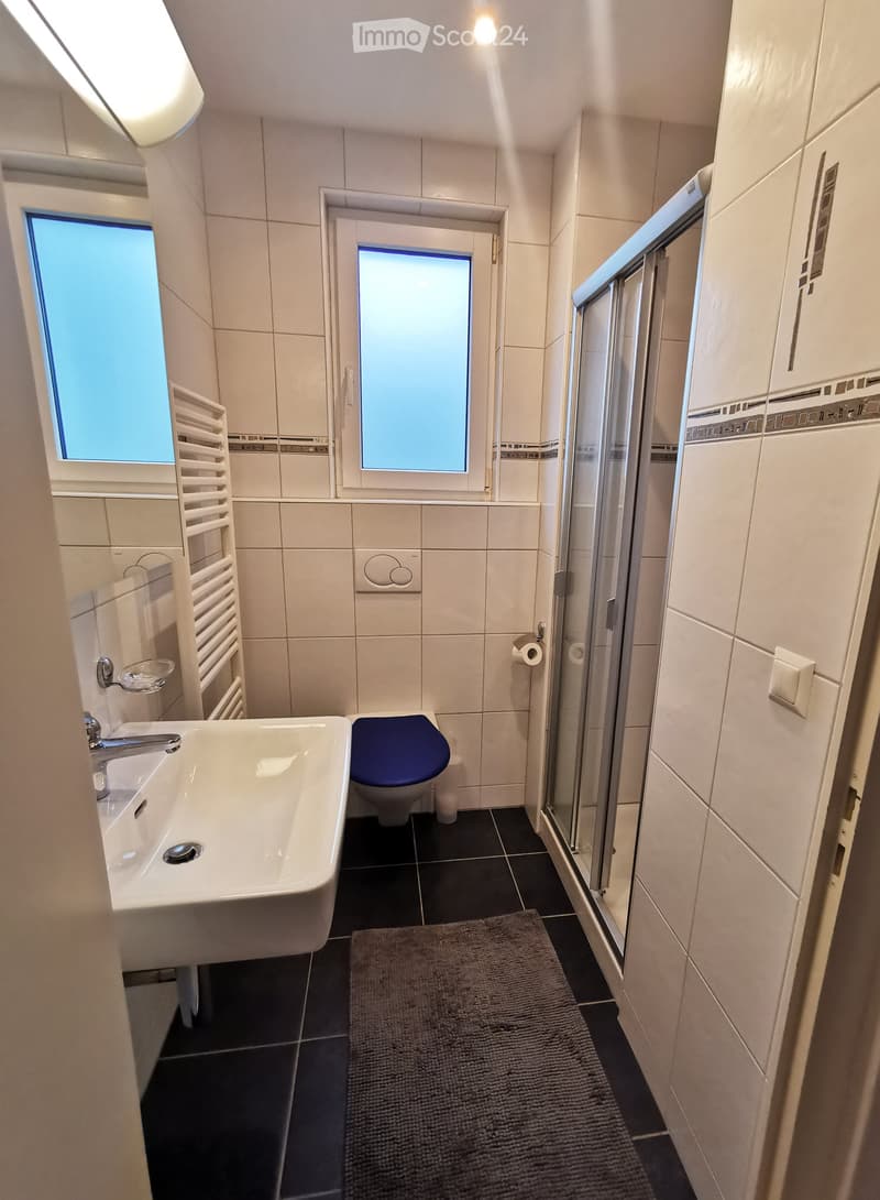 Modern bathroom with white tiles, a bathtub, toilet, sink, and a glass-enclosed shower. Features a towel rack and a small rug on the floor.