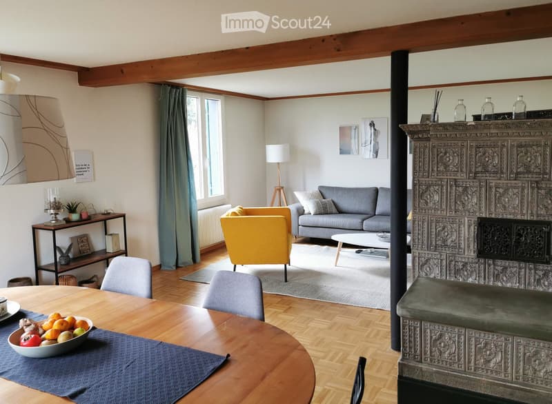 Living room with wooden flooring, exposed wooden beams, grey sofa, yellow armchair, coffee table, dining table with chairs, fireplace, shelves, and decorative items.