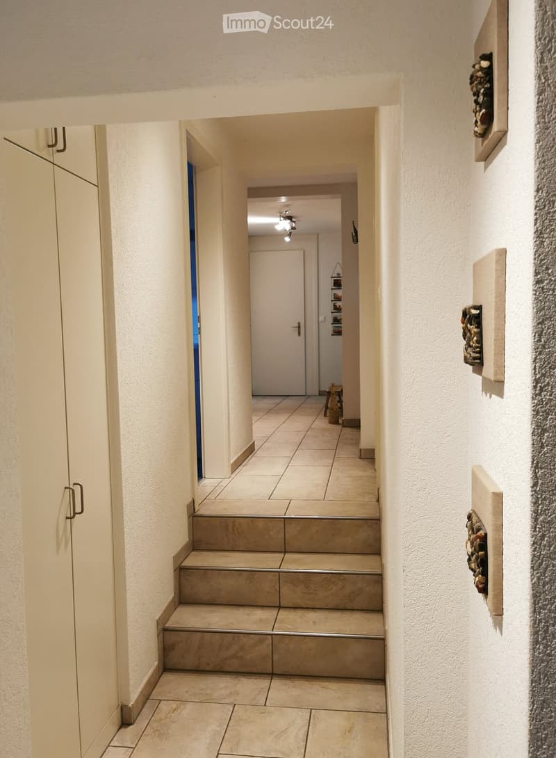 A clean, modern hallway with tiled flooring, white walls, and a staircase leading upwards. The hallway features a closed door on the right and a partially open door on the left, with decorative wall pieces visible on the right side.