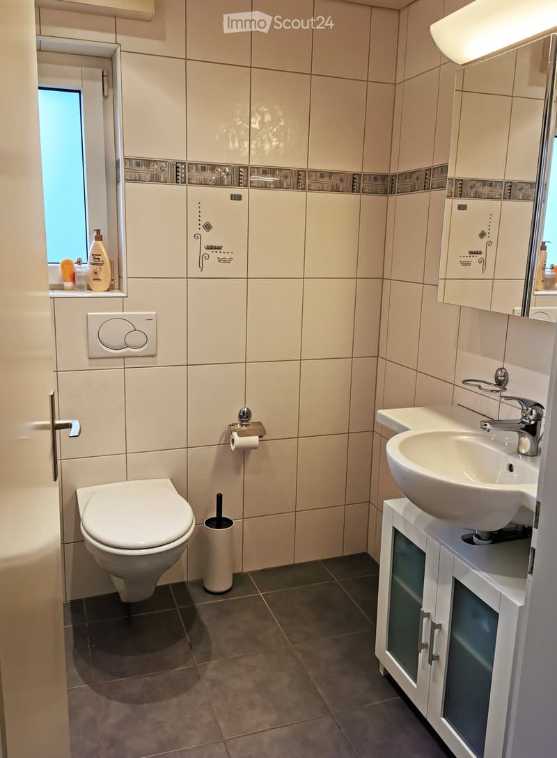 Modern bathroom with white tiles, a toilet, a sink with a modern faucet, a cabinet, and a window providing natural light.