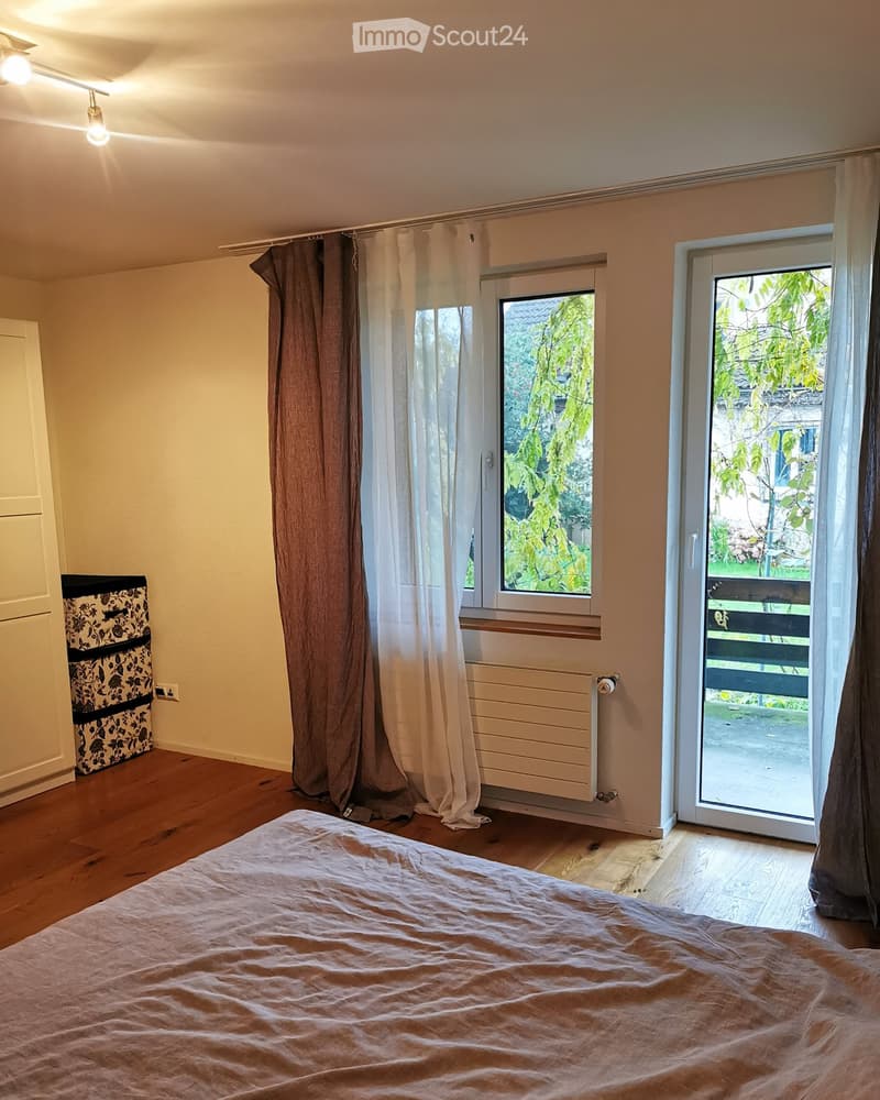 Bright bedroom with wooden flooring, large window with curtains, sliding glass door leading to a balcony, and a neatly made bed.
