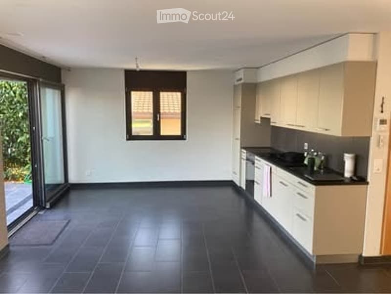 Open-plan kitchen and living area with white walls, dark tiled floor, built-in kitchen cabinets, and a large window with blinds. The space is completely empty, with no furniture or appliances visible.