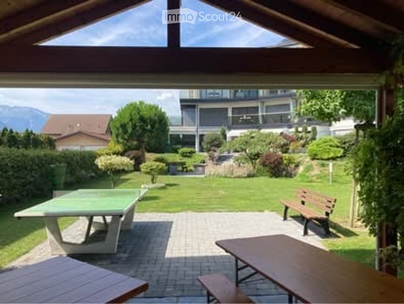 Well-maintained garden with a ping pong table, wooden benches, and outdoor furniture, surrounded by greenery and a view of mountains.