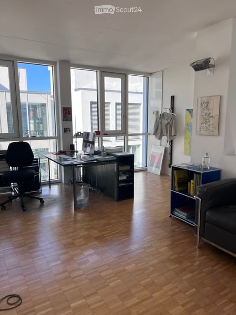 Modern office space with large windows, wooden flooring, a desk with a laptop, a chair, a bookshelf, and a small sofa.